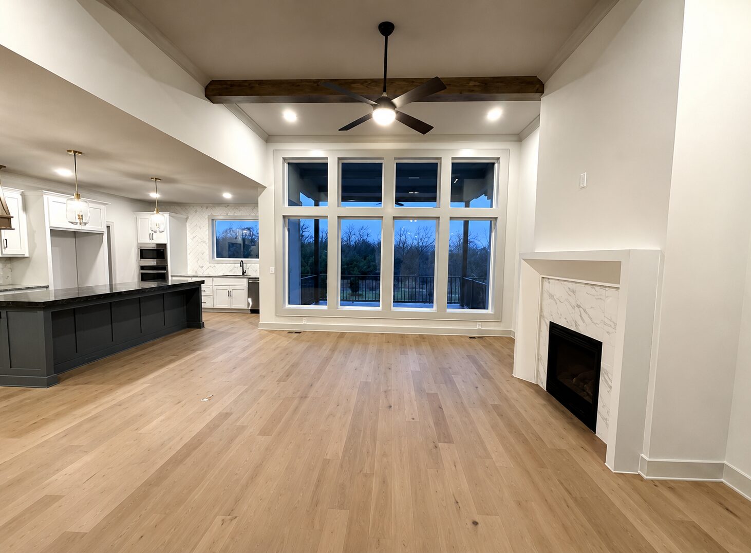 Spacious custom home great room with floor-to-ceiling windows, exposed wood ceiling beams, hardwood floors, a modern fireplace, and an open kitchen with a large island.